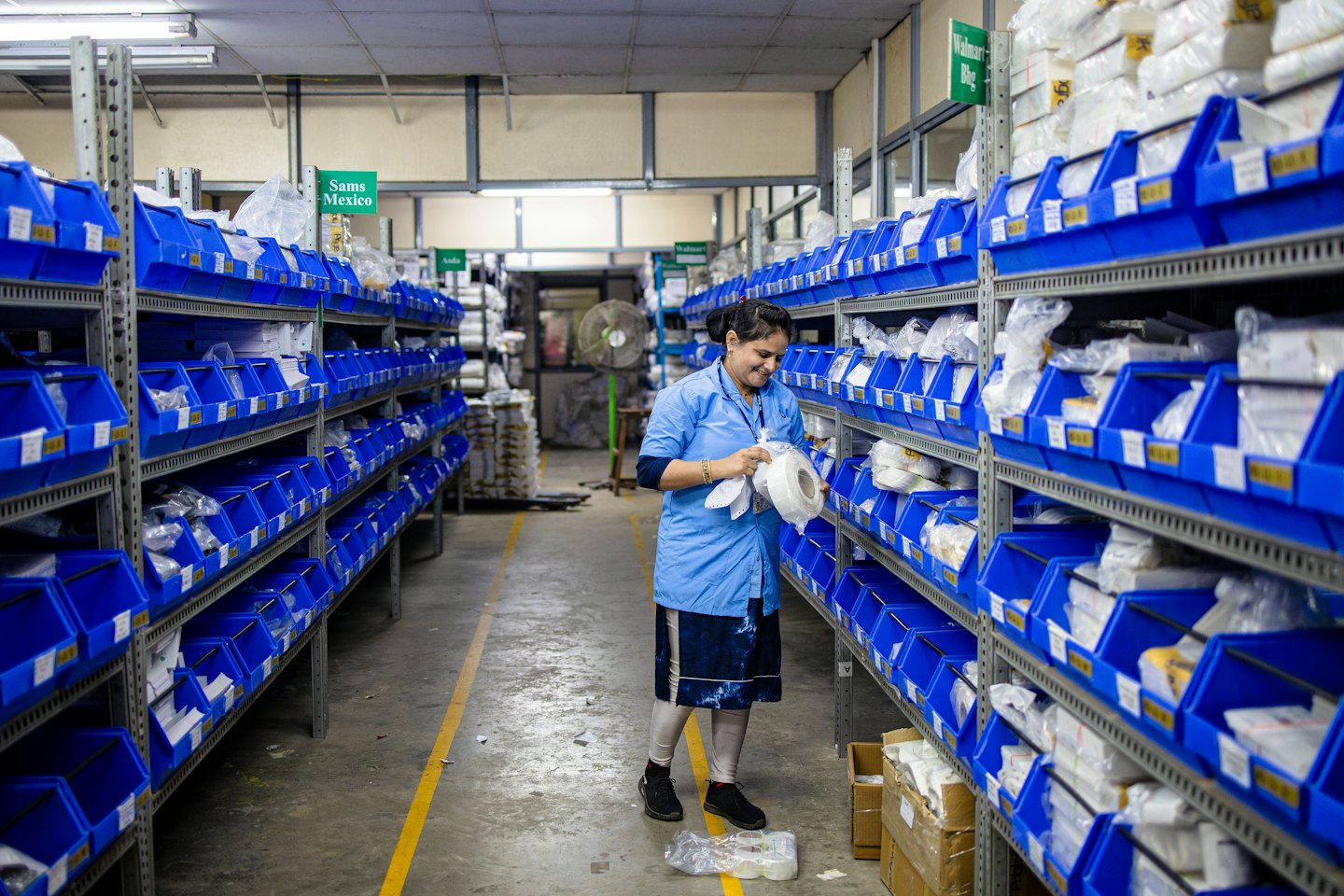 Sieviete zilā uniformā šķiro preces no plastmasas tvertnēm noliktavā.